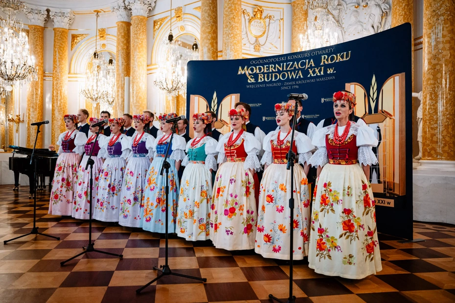 Zespół w tradycyjnych strojach ludowych występuje na scenie podczas uroczystej gali wręczenia nagród konkursu „Modernizacja Roku & Budowa XXI w.” na Zamku Królewskim w Warszawie.Występ zespołu folkowego w kolorowych strojach ludowych na tle ścianki konkursu Modernizacja Roku i Budowa XXI w. na Zamku Królewskim.